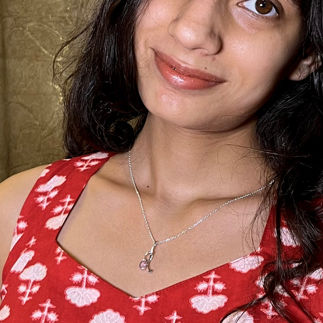 A close-up lifestyle shot of a woman wearing a delicate silver necklace with a small pink stone pendant, paired with a red and white patterned top.